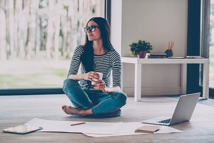 woman sitting on the floor, drinking a cup of coffee, and looking at sheets laid out on the floor