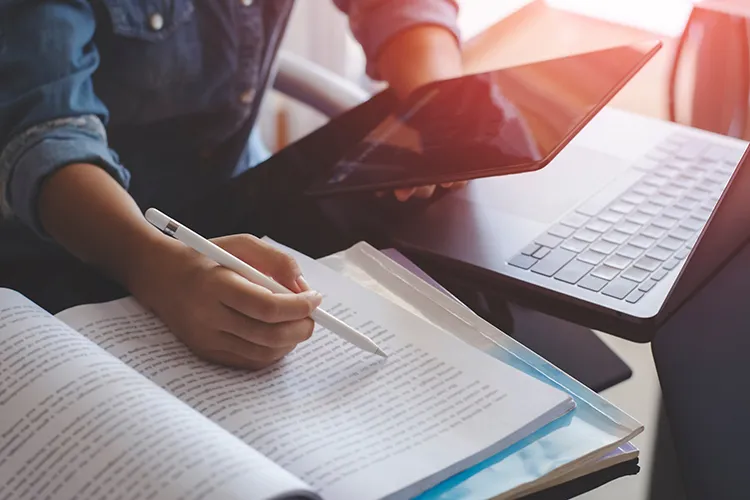 student using a laptop, textbook, and tablet to complete school work